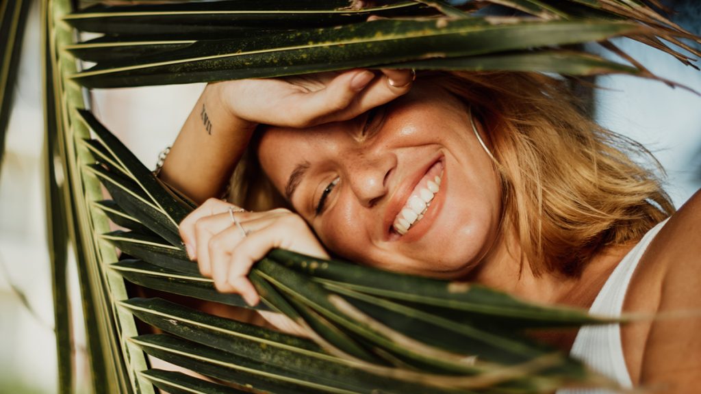 A joyful woman smiling through palm leaves, with sunlight highlighting her face, visible tattoos, and tousled hair.
