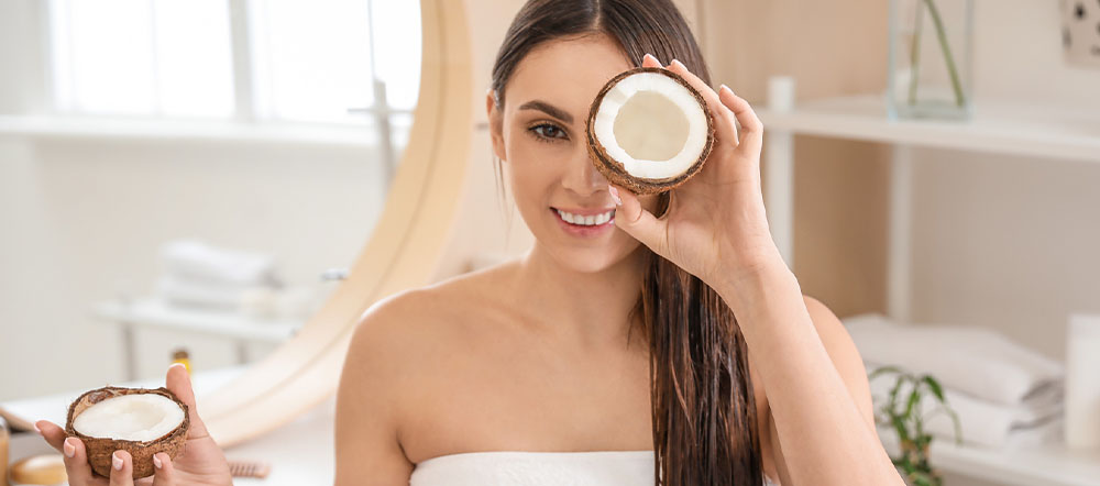 A smiling woman holding a halved coconut over one eye, sitting in a well-lit spa-like setting.