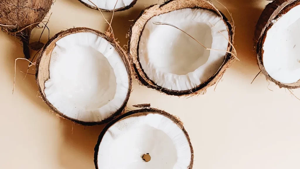 Several halved coconuts with their white flesh exposed are arranged on a light surface. The rough, fibrous brown shells are partially intact, with some coconut water residue visible inside a few halves.