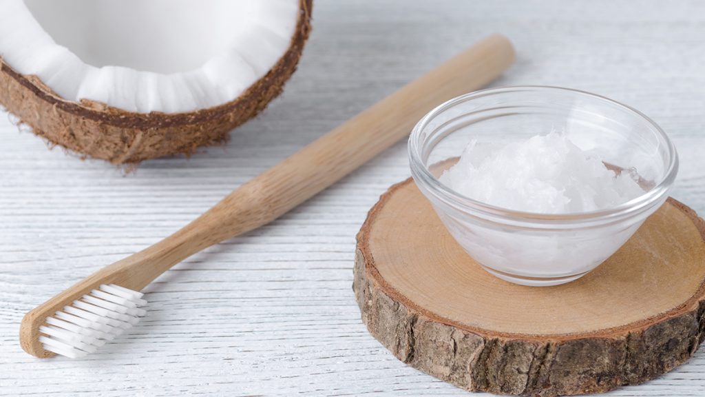 A wooden toothbrush beside a glass bowl of coconut oil, set on a wooden disc, with a halved coconut in the background on a white wooden surface.