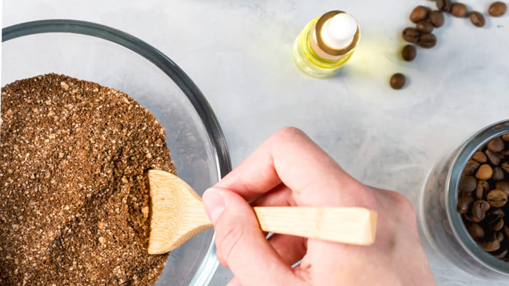 Close-up of a person's hand spreading a coffee ground scrub mix into a glass bowl using a wooden spatula, with a bottle of oil and whole coffee beans nearby on a marble surface.