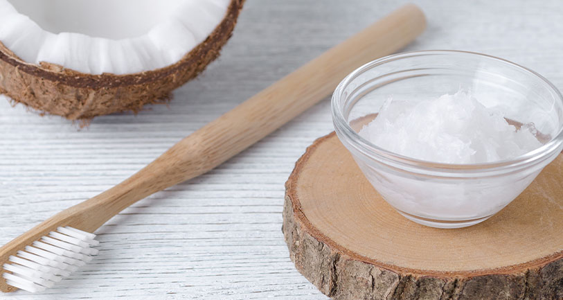 A wooden toothbrush and a clear bowl filled with coconut oil sit on a wooden slab, with a halved coconut in the background on a white wooden surface.
