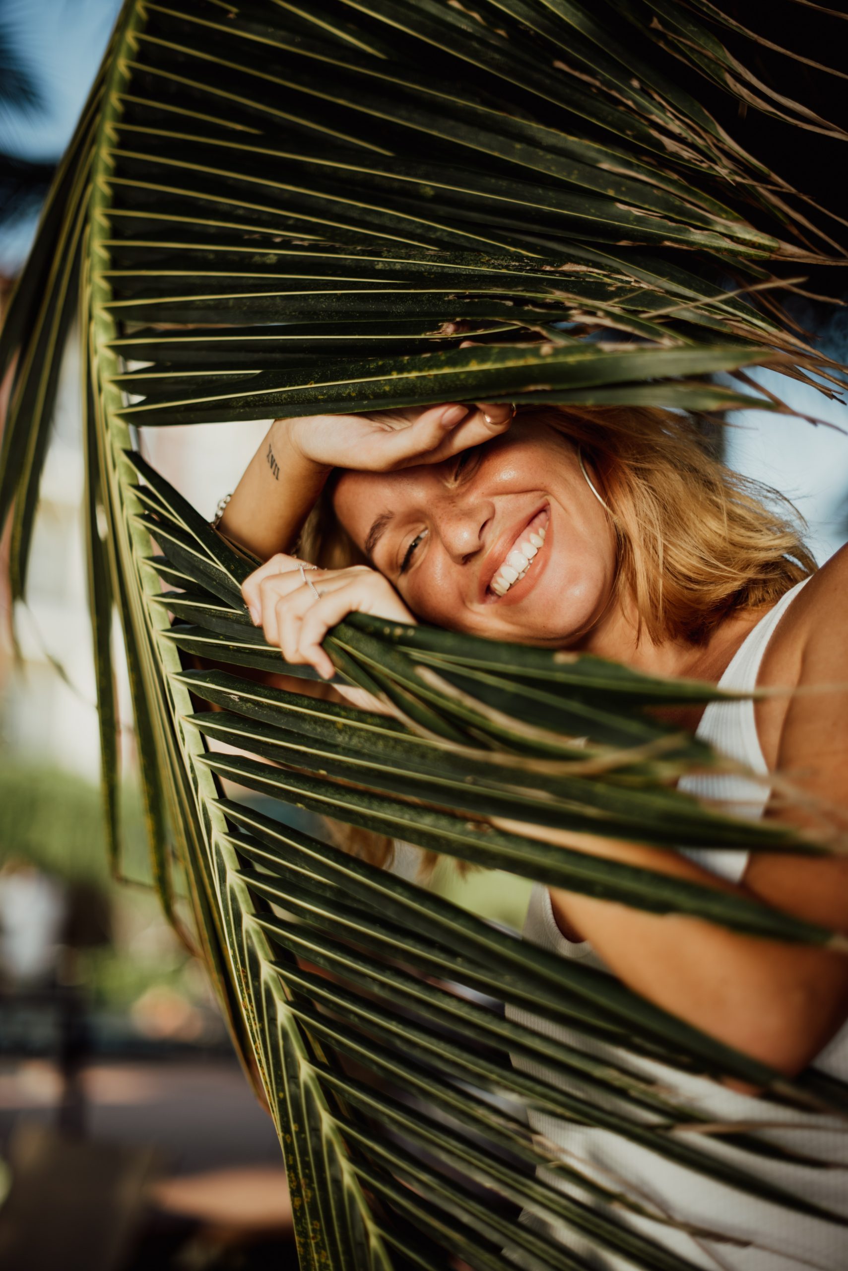 A joyful woman peeks through large palm leaves, smiling broadly and covering her forehead with her hand, immersed in a sunny, tropical setting.