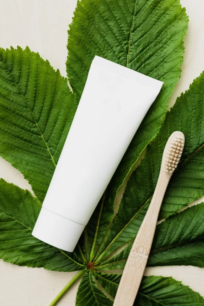 A white toothpaste tube and a bamboo toothbrush are placed on a large green leaf. The leaf serves as a natural background, enhancing the eco-friendly theme suggested by the bamboo toothbrush.
