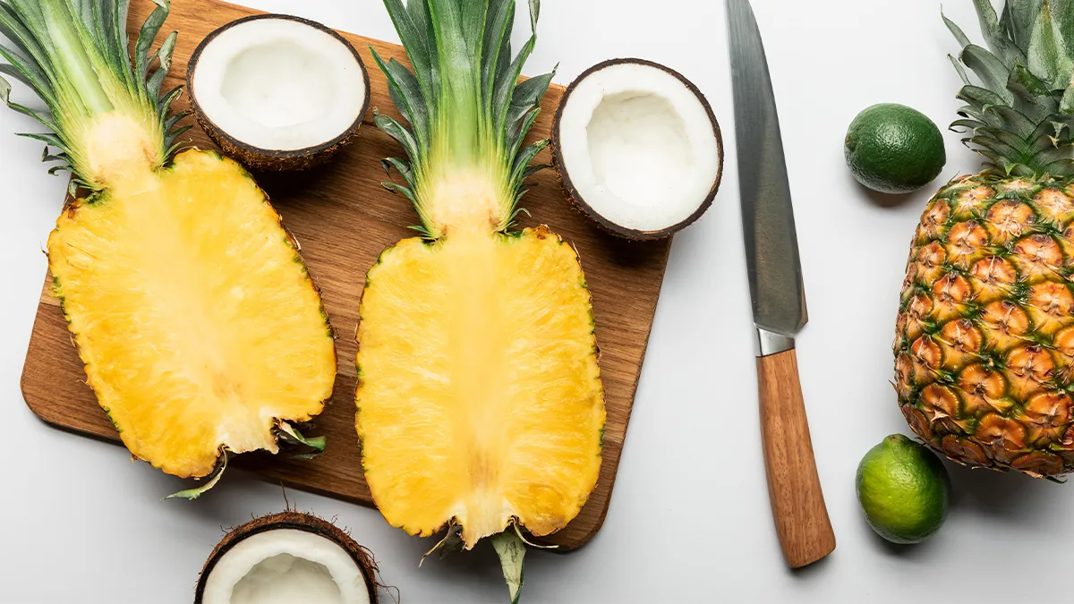 Two pineapple halves on a wooden cutting board, surrounded by three coconut halves, a whole pineapple, two limes, and a knife with a wooden handle on a white surface. The fruit arrangement displays vibrant and fresh tropical produce.