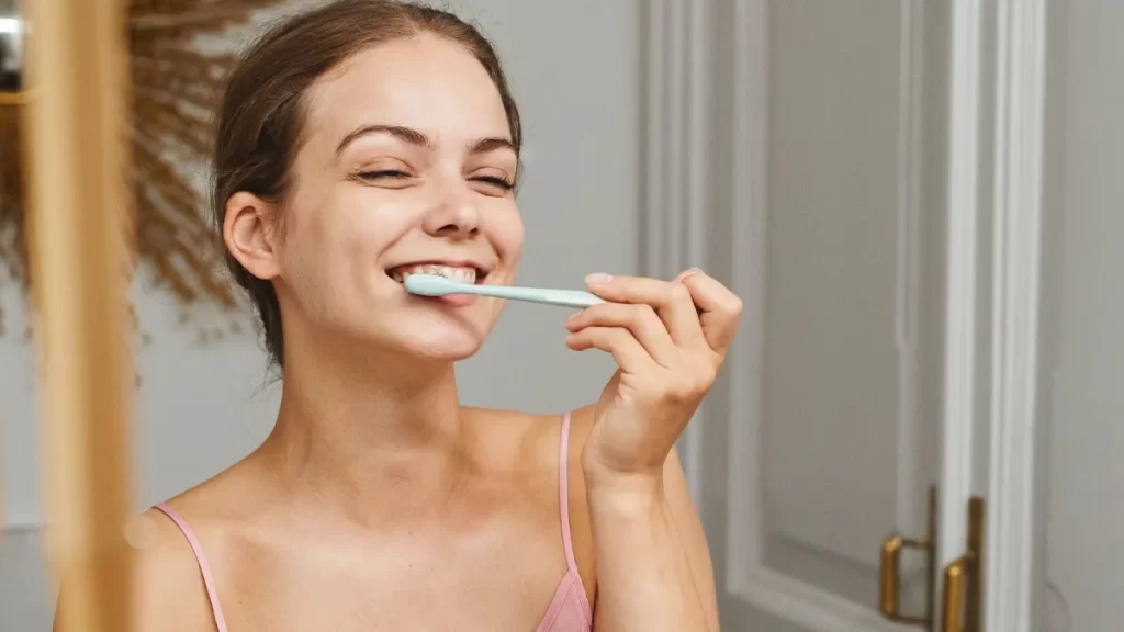 A young woman is smiling with her eyes closed while brushing her teeth with a light blue toothbrush. She is wearing a pink tank top and standing in a bathroom with light-colored walls.