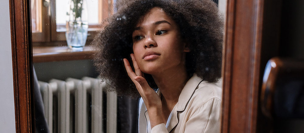 A young woman with curly hair reflects thoughtfully in a mirror, touching her face, in a warmly lit room.