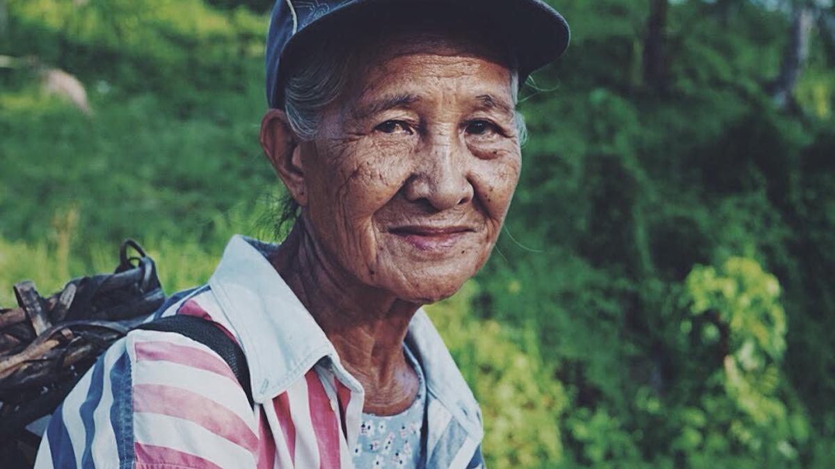 An elderly woman with a content smile, wearing a cap and striped shirt, carries a bundle of sticks on her back, with lush greenery in the background.