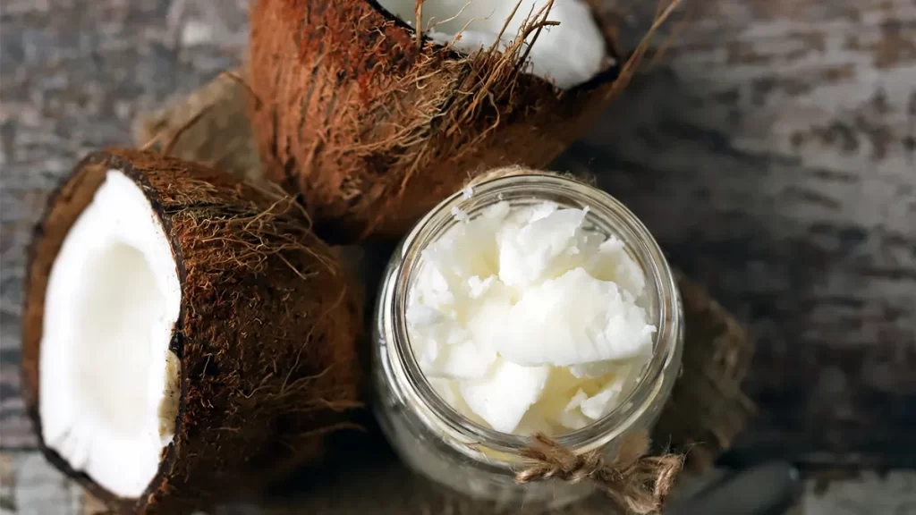 A jar of coconut oil sits on a rustic wooden surface, surrounded by two fresh, halved coconuts. The oil in the jar appears solid, and the texture of the coconut shells is rough and fibrous.