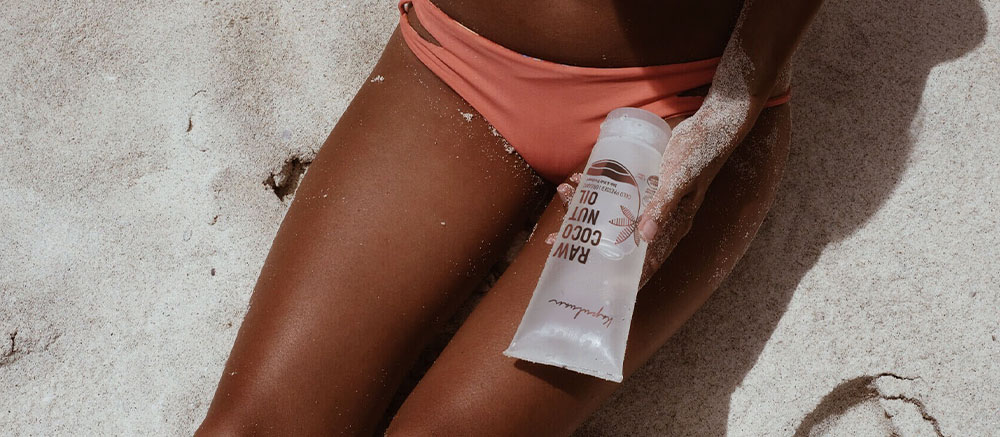 A close-up of a person's sun-kissed legs on a sandy beach, holding a tube of sunscreen labeled "bare republic.