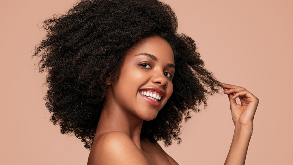 A joyful young woman with voluminous curly hair, smiling broadly against a soft pink background, playfully touching her hair.