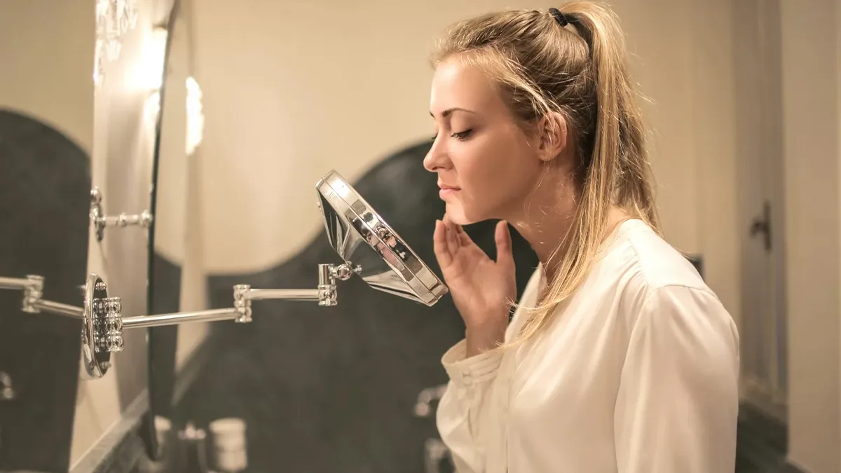 A woman with blonde hair tied back in a ponytail is examining her face in a small round magnifying mirror attached to the wall in a bathroom. She is wearing a white top and appears to be focusing on her complexion.