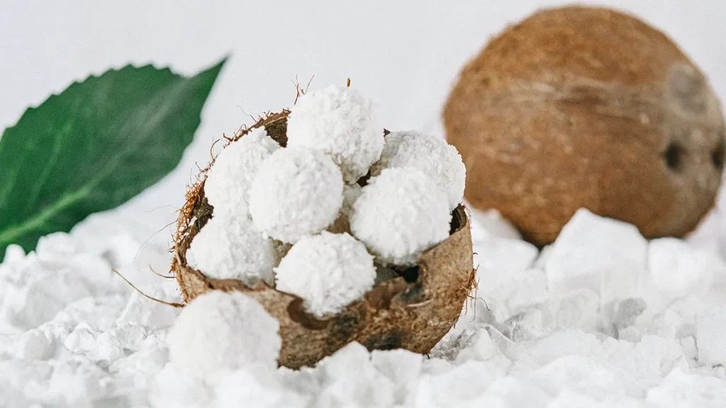 Coconut candies displayed inside a halved coconut shell, surrounded by white stones, with a whole coconut and green leaf in the background. The candies are spherical and coated in white, resembling coconut flakes.