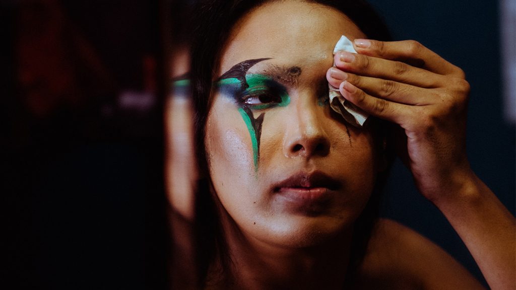 A woman removes vibrant green stage makeup from her face using a tissue, with a focus on her eyes, partly covered in striking makeup.