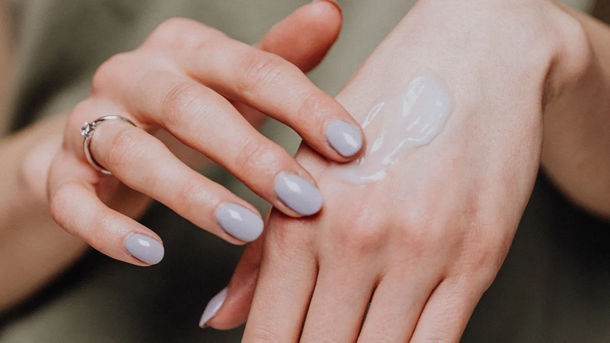 Close-up of a person applying a dollop of white lotion on the back of their hand with their other hand. The person has neatly manicured nails painted in a light gray color and is wearing a silver ring on one finger. The background is blurred and out of focus.