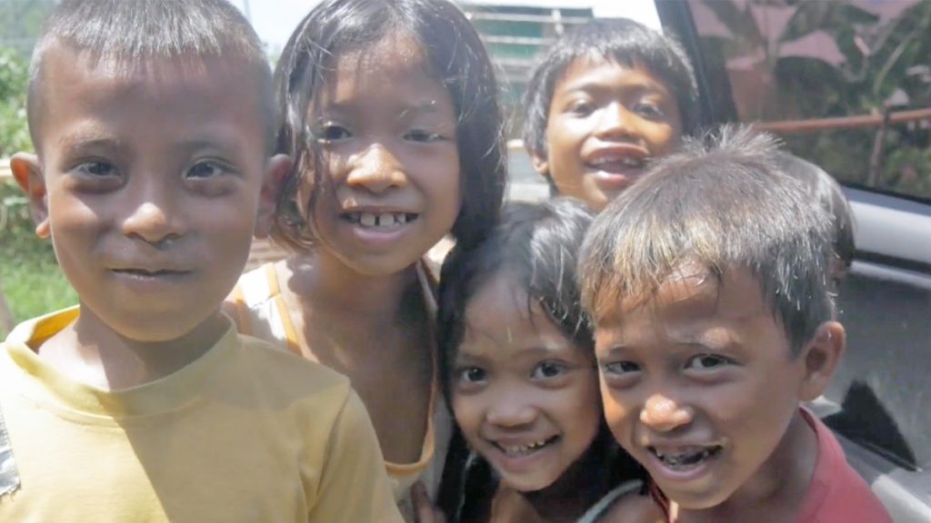 Six joyful children with bright smiles gather closely for a group photo outdoors on a sunny day.