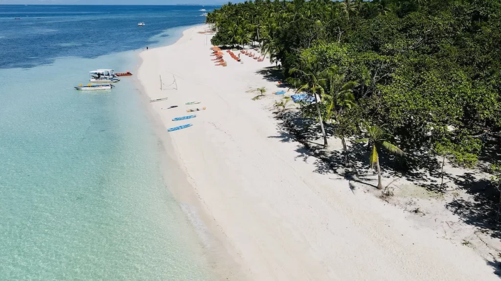 Aerial view of a serene beach with white sand and clear turquoise water. Palm trees and greenery line the shore. Several kayaks, sun loungers, and umbrellas are spread out on the beach. Boats are anchored in the shallow waters. The scene is tranquil and idyllic.