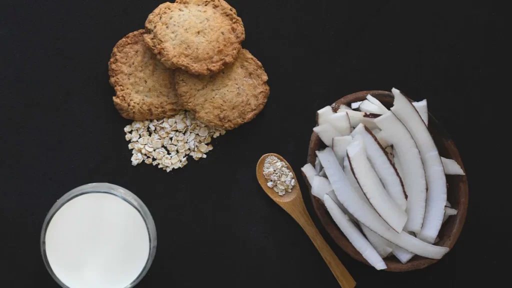 A glass of milk, a bowl of freshly cut coconut pieces, three oatmeal cookies, and a small pile of oats are arranged on a black surface. A wooden spoon containing oats is placed beside the bowl of coconut pieces.