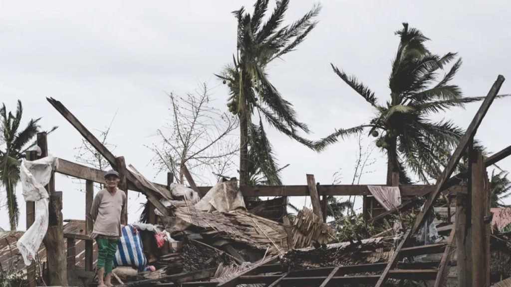 A person stands amid the wreckage of a destroyed home with debris scattered around, surrounded by storm-damaged palm trees, conveying the aftermath of a natural disaster.