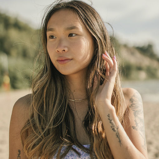 A woman with long hair and a floral tattoo on her arm stands on a beach, touching her hair and gazing off to the side.