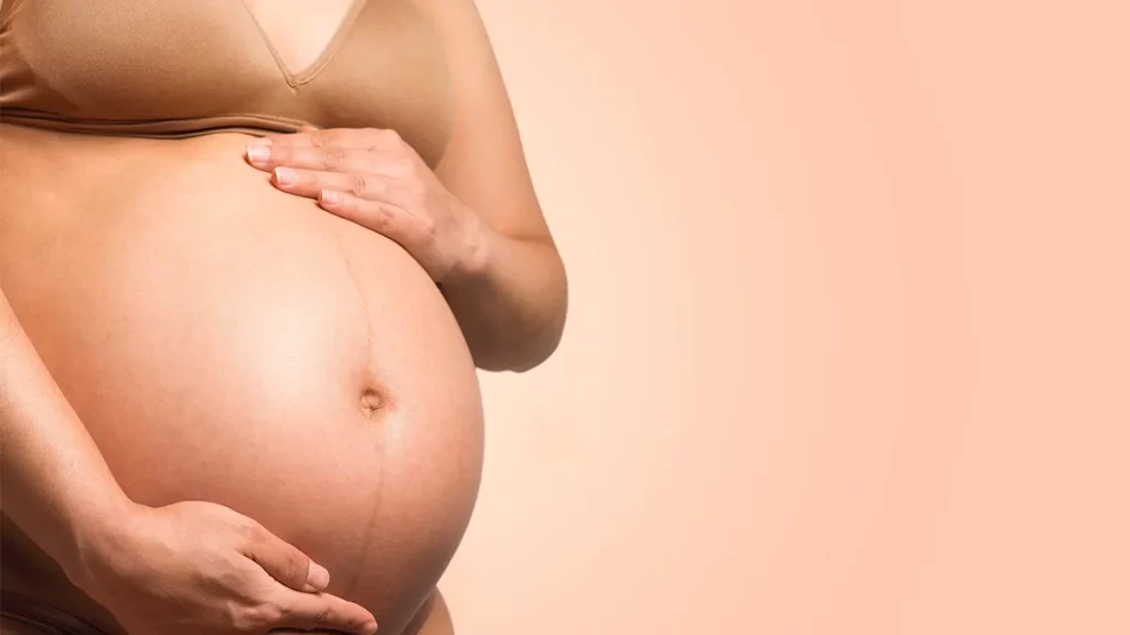 A close-up image of a pregnant person holding their bare, round belly with both hands, one on top and one below. The background is a soft peach color.