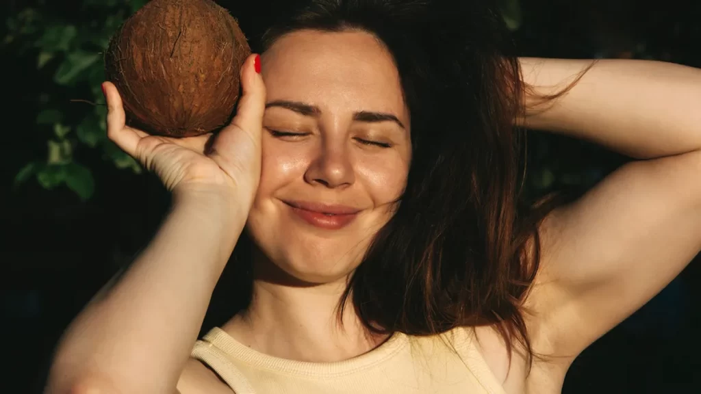 A woman with long brown hair smiles with her eyes closed, holding a coconut near the left side of her face. She has one hand behind her head, and sunlight illuminates her face and arm. She wears a beige tank top and has red nail polish on her fingernails.