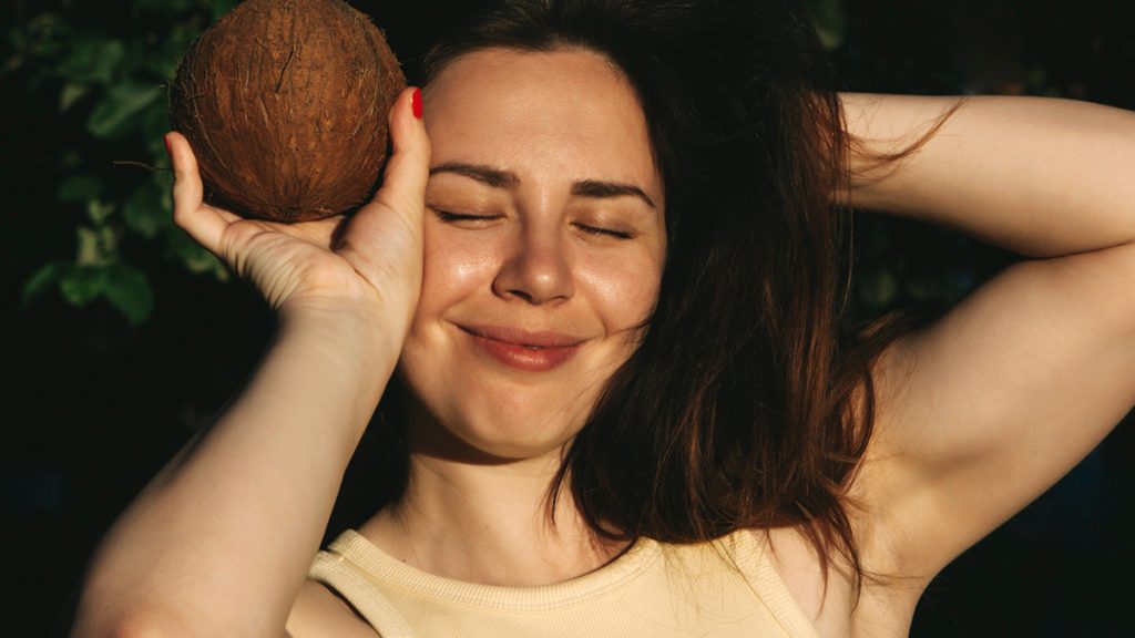 A woman with a joyful expression holds a coconut near her face, enjoying the sun in a wooded area. her eyes are closed, and she has a relaxed smile.