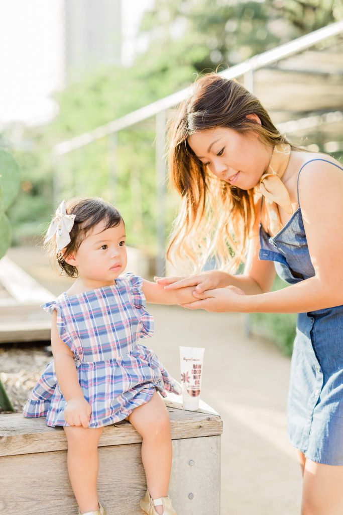 A young woman applies sunscreen on a toddler girl's arm outdoors on a sunny day. both are dressed in summer attire, with the child looking curiously at the woman.