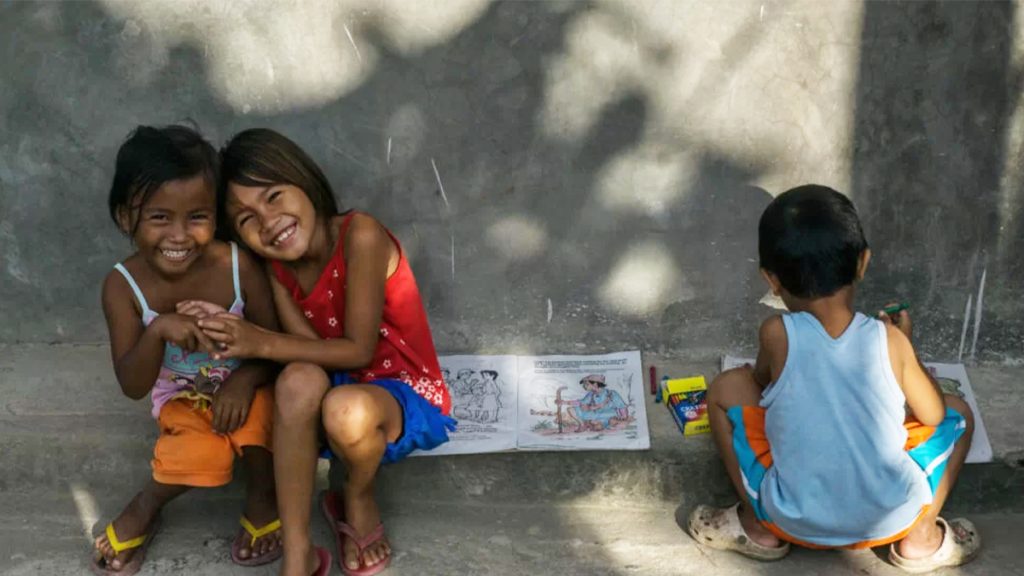 Two joyful children, one girl hugging another, smile brightly while sitting on a sidewalk. another child in a blue shirt plays nearby. a drawing is visible between them on the ground.