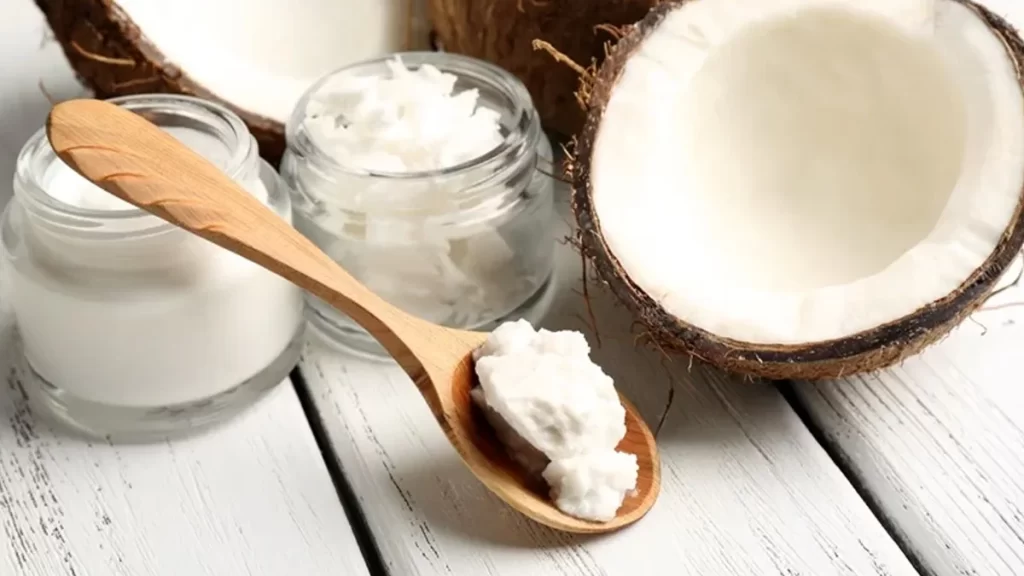 A wooden spoon with coconut butter resting on it is placed next to a split coconut, a small open jar of coconut cream, and a jar of coconut flakes. All items are arranged on a white wooden surface.