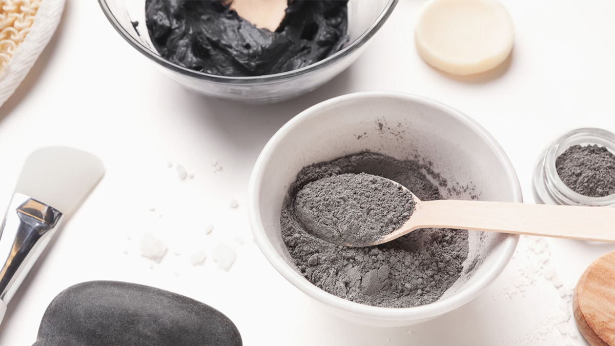 Beauty and skincare products displayed on a white surface, including bowls of black clay mask and powder, a brush, a soap, and various applicators.