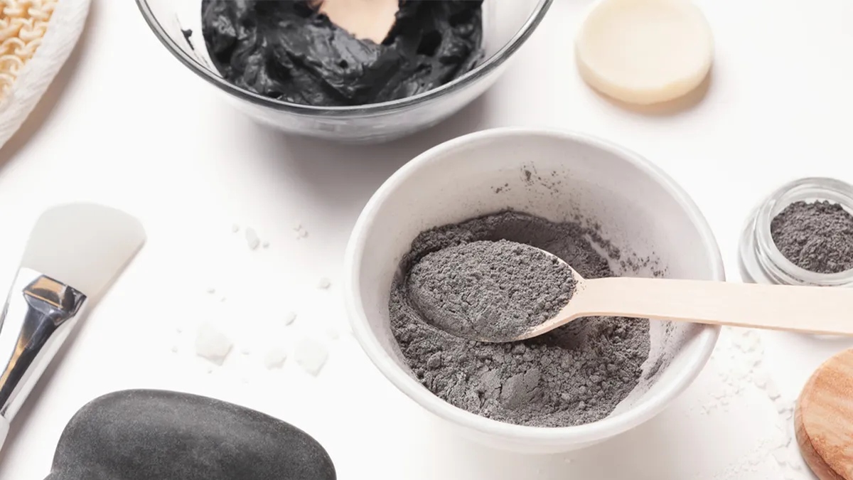 A white bowl filled with gray powder and a wooden spoon placed inside. In the background, there is a bowl with black paste and various skincare tools and products, including a brush, a small jar, and a black stone, all arranged on a white surface.