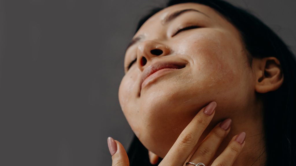 Close-up of a smiling asian woman with her eyes closed, touching her face gently, highlighting her clear skin against a gray background.