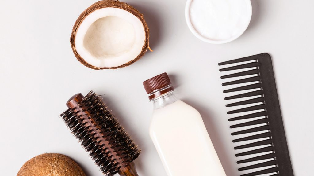 Overhead image of coconut halves, a bottle of coconut milk, natural hair brushes, and a comb on a pale background, depicting hair care products.
