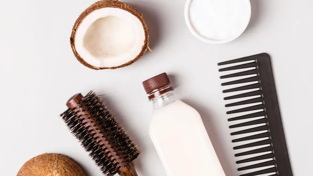 Top view of hair care items including a brown round brush, black wide-tooth comb, bottle of white liquid with brown cap, halved coconut, and small white bowl of creamy substance, all arranged on a white background.
