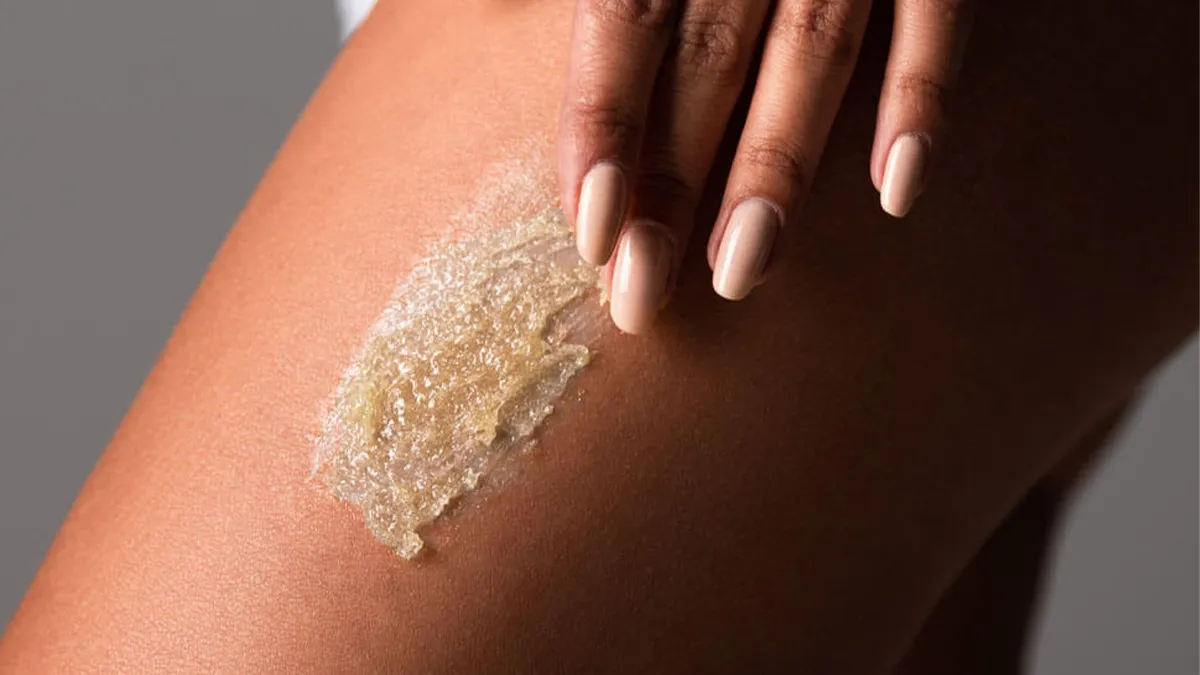 Close-up of a person applying a body scrub to their thigh. The person has medium to dark skin tone and well-manicured, nude-colored nails. The image captures the texture of the scrub, highlighting its granulated consistency.