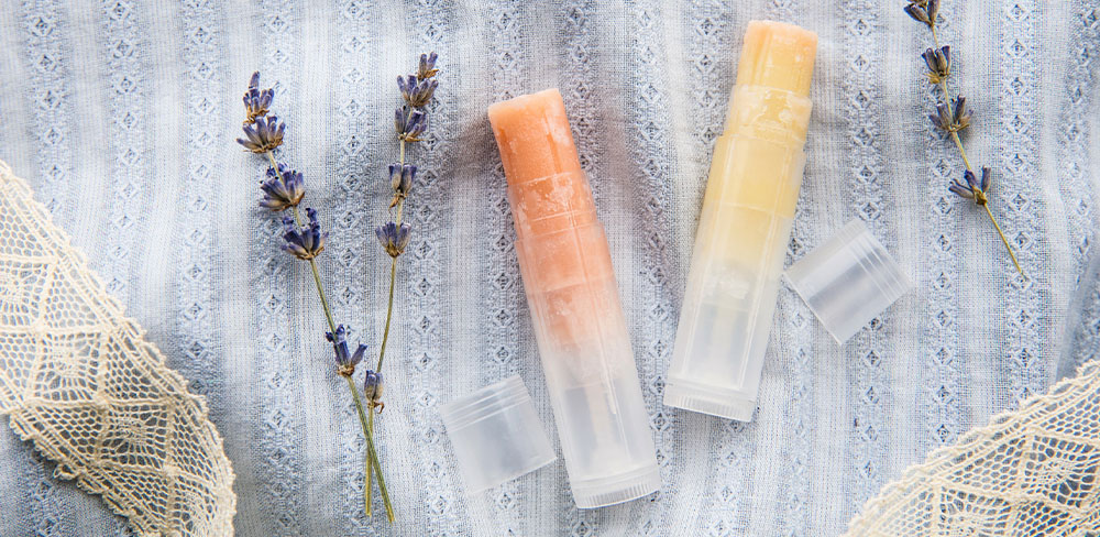 Two lip balms in clear tubes alongside sprigs of lavender, placed on a lace-covered light fabric. the image exudes a natural and soothing aesthetic.