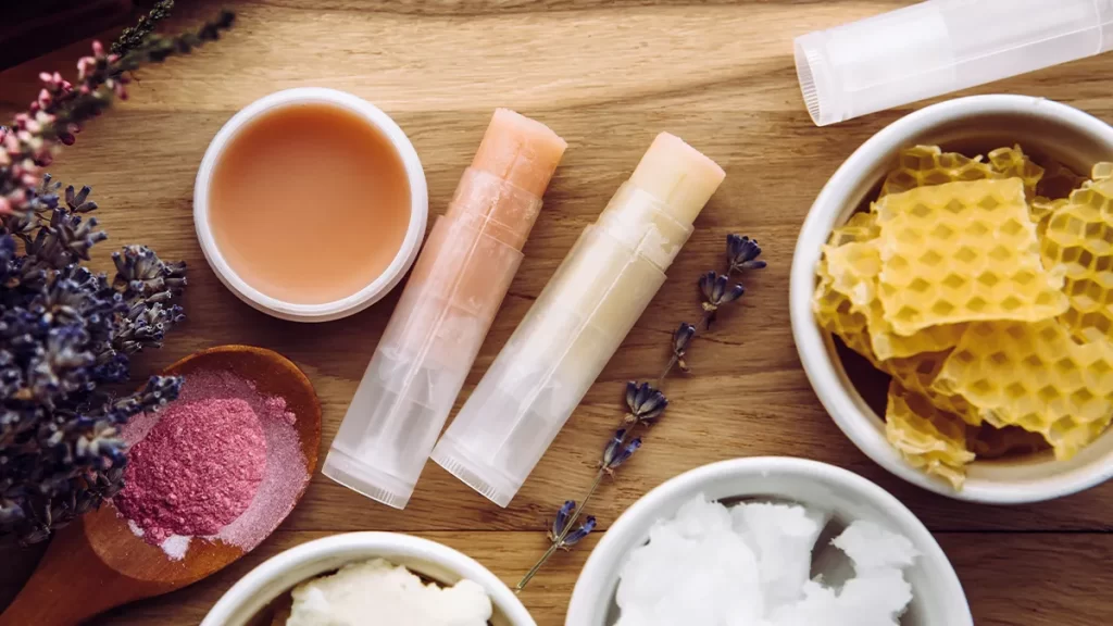 A collection of natural lip balm ingredients on a wooden surface. Two lip balm tubes, a small jar of balm, a bowl with beeswax chunks, a wooden spoon with pink powder, and a bowl with white solid substance. Sprigs of dried lavender are scattered around.
