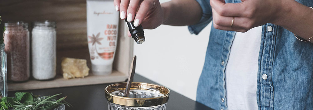 A person in a denim shirt adding a liquid ingredient to a bowl in a kitchen, with herbs and spices visible on the counter.