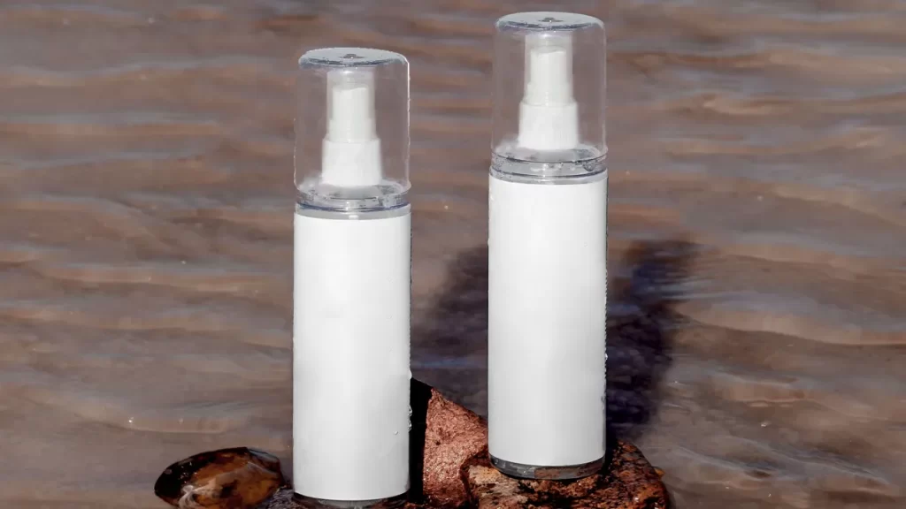 Two white cylindrical spray bottles with clear caps are standing upright on a wet rock, surrounded by shallow water. The spray bottles are identical in design, with one being slightly smaller than the other.