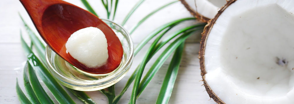 A wooden spoon holds a round white coconut oil ball above a glass dish, surrounded by vibrant green palm leaves and a halved coconut on a white wooden surface.