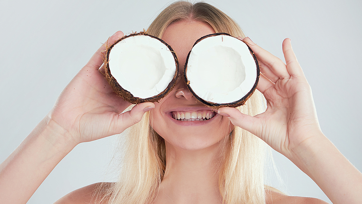 A smiling woman holds two halves of a coconut in front of her eyes like binoculars, against a light gray background.