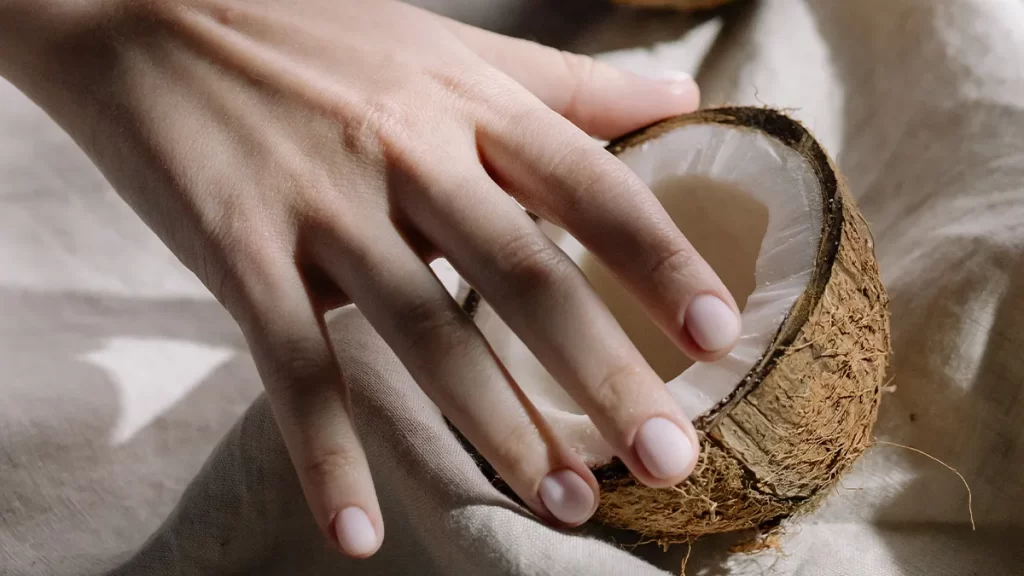 A hand gently rests on a halved coconut, with the fingers touching the inner white flesh. The coconut is placed on a fabric surface, and the scene is softly lit.