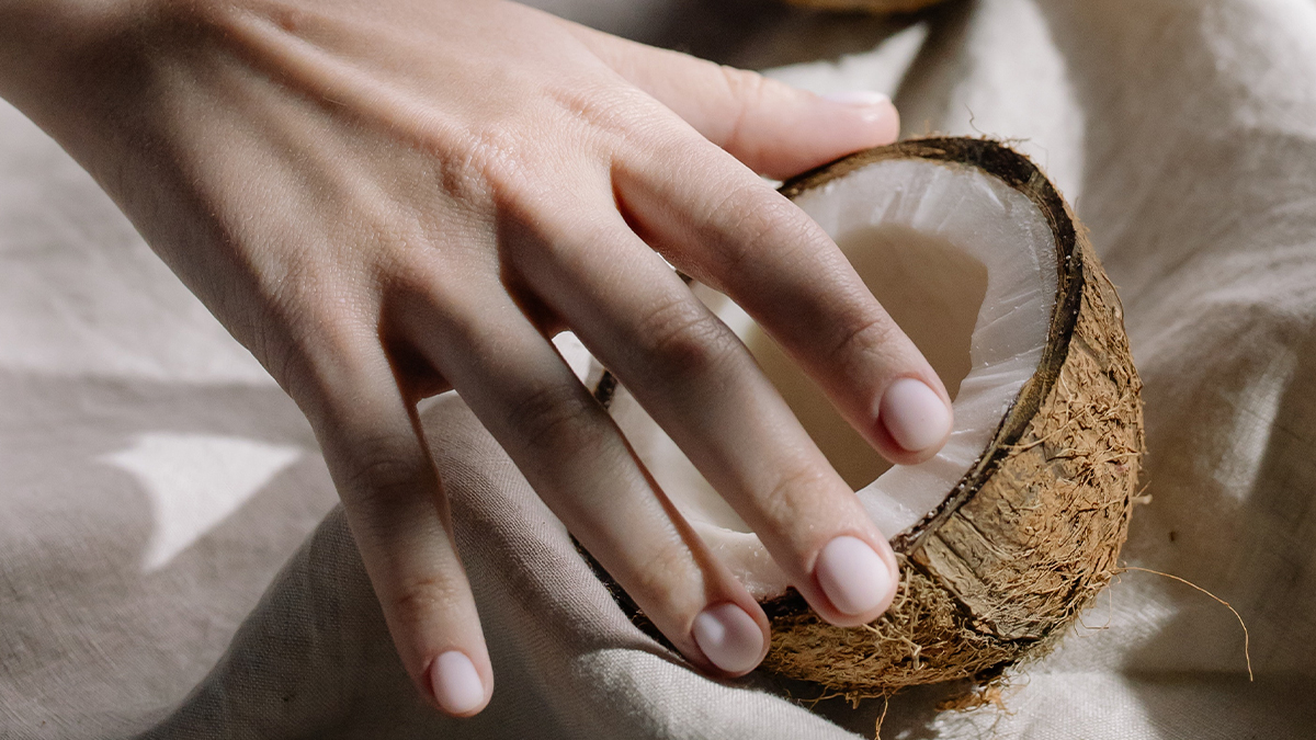 A person's hand with manicured nails gently holding a half coconut shell on a textured fabric under natural light.