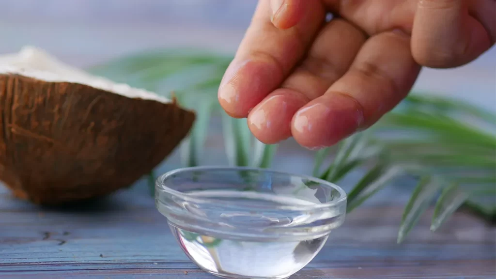 A hand with fingers slightly touching and glistening, held above a small glass bowl filled with clear liquid. A halved coconut and green leaves are blurred in the background, all resting on a blue wooden surface.