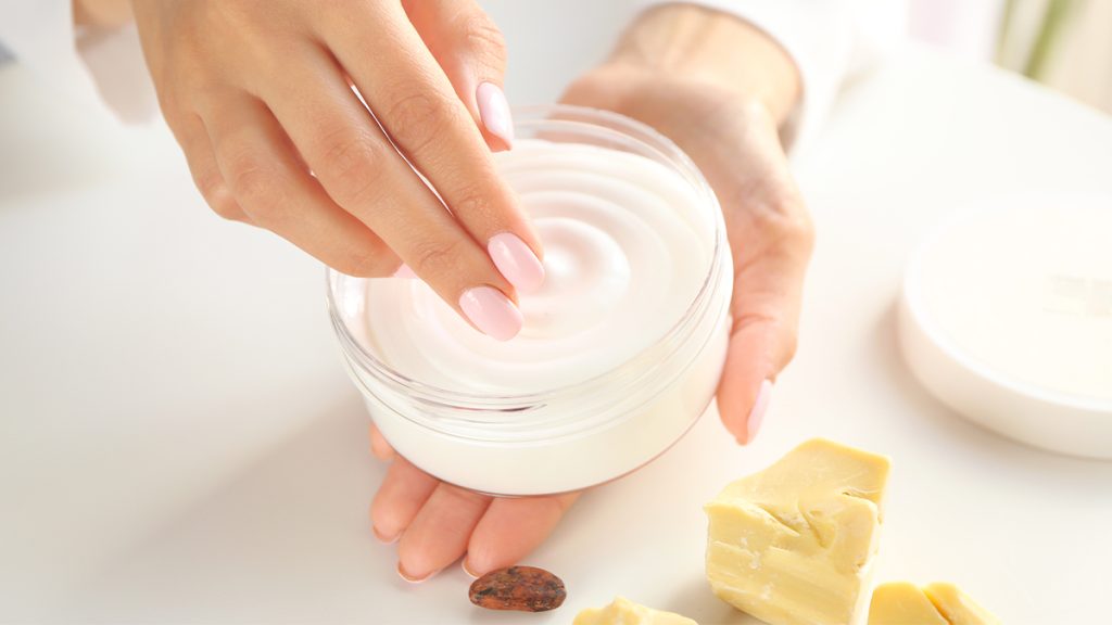 A person's hands delicately touching a jar of white cream, with blocks of natural soap and a cocoa bean nearby on a light surface.