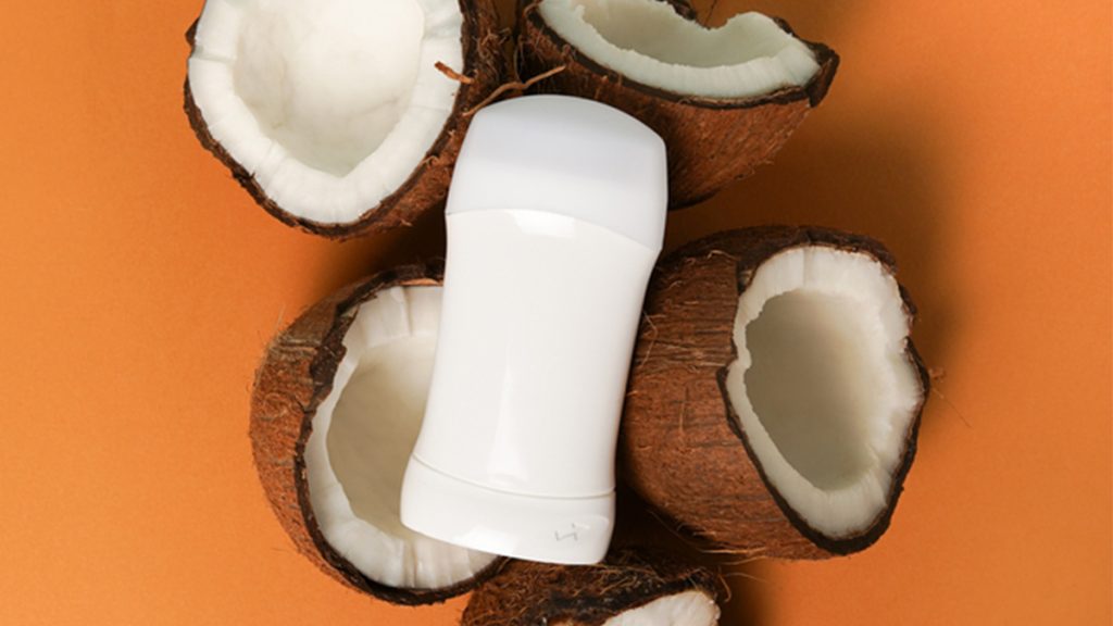 A deodorant stick placed among several half-shells of fresh coconuts on an orange background, suggesting a coconut-scented product.