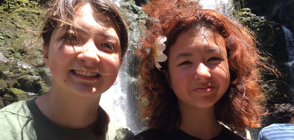 Two young women smiling in front of a waterfall, one with a flower in her hair.