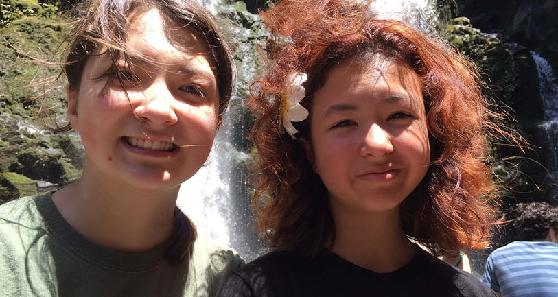 Two smiling young women standing in front of a waterfall. one has a flower in her hair. they are outdoors on a sunny day, surrounded by lush greenery.
