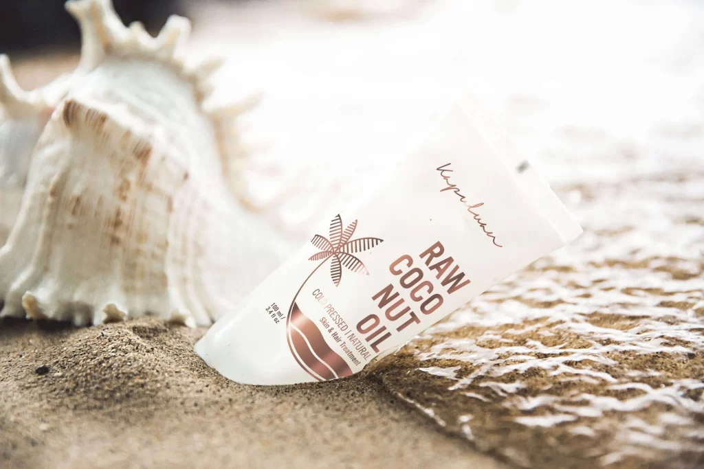 A tube of raw coconut oil by Kopari sits on sandy ground next to a large seashell. The tube is white with brown text and a small illustration of a palm tree. The background shows a soft focus of what appears to be a beach with gentle waves.